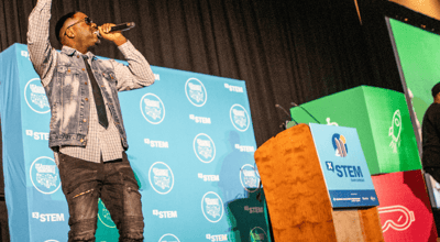 An African American man in a shirt, tie and demin jacket speaks on stage during an X-STEM event.